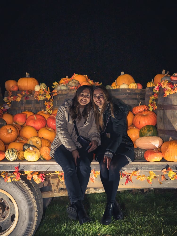 2 ladies sat in between a bunch of pumpkins and barrels at the patch mk